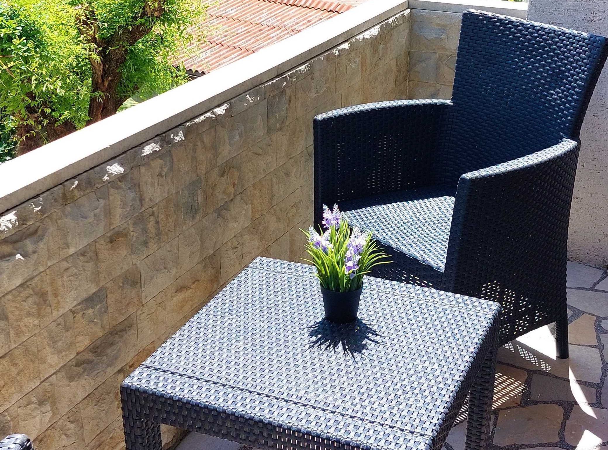 A black wicker chair and a small square table with a woven tabletop, placed on a stone patio with a stone wall and greenery in the background.