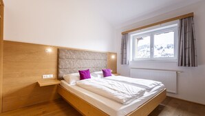 A modern bedroom with a wooden headboard, a large window with curtains, and a bed with white bedding and purple pillows.