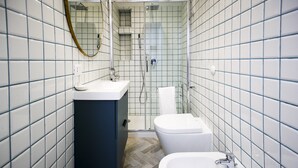 Un baño moderno con paredes de azulejos blancos, suelo de madera oscura, lavamanos blanco, inodoro y ducha con mampara de cristal.