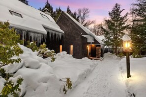 Une cabane en rondins dont le toit est recouvert de neige et des glaçons pendent des avant-toits, le tout étant entouré de neige et d’arbres.