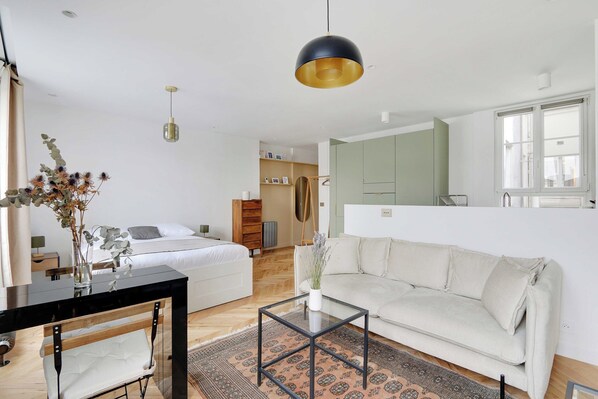 A modern living room with a sofa, a coffee table, a dining table, and a hanging light fixture. - Charming Studio - 2P - Canal (Paris)