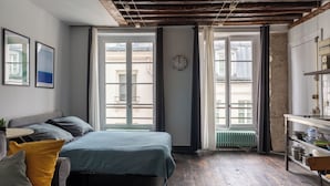 A room with a bed, a small table, a radiator, and a window with curtains. - Le Marais St.Paul Mobility lease apartment  (Paris)