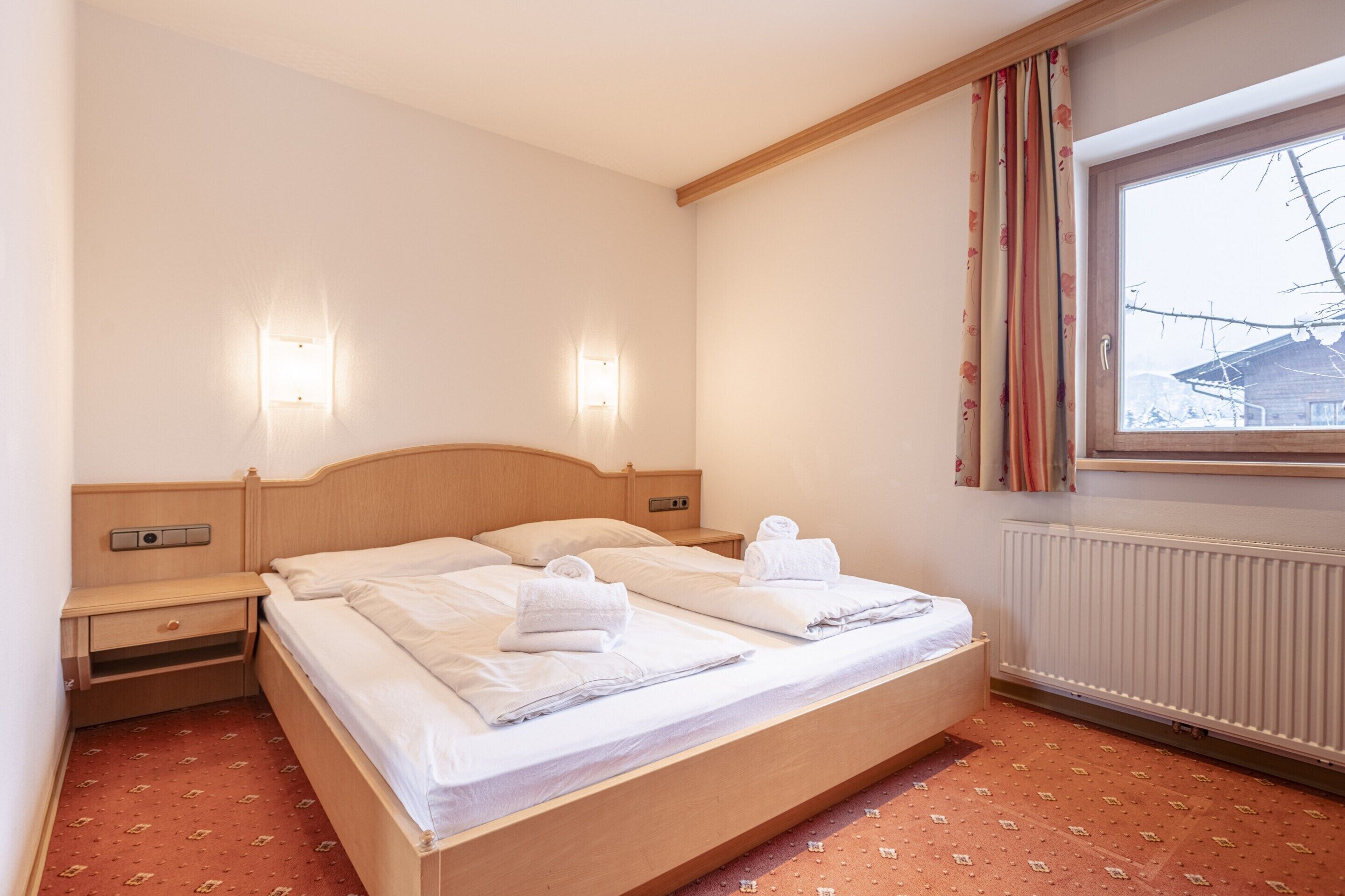 Une chambre d’hôtel avec un lit double, deux tables de chevet, une petite table et une fenêtre avec des rideaux.