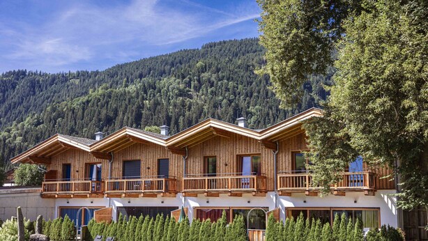 A wooden chalet with balconies, surrounded by a well-maintained lawn and trees, set against a mountainous backdrop.