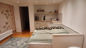 A bedroom with a bed, a small table, a radiator, a wooden floor, and a kitchen area in the background. - Beautiful vacation room near Wutachschlucht (Löffingen)