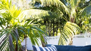 A dark green outdoor sofa with a striped pillow, surrounded by palm trees and a white fence. - *NEW* Riverside Retreat w/ Pool (Jupiter)