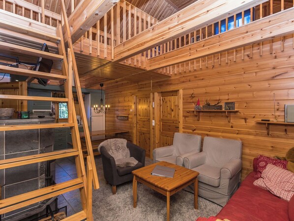 A cabin interior with a loft bedroom, a living area with a sofa and armchairs, a dining table, and a kitchen area.