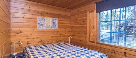 A wooden cabin room with a bed, a small table, a lamp, and a window with a view of trees.