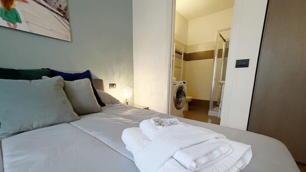 A neatly made bed with white linens and a folded towel, a nightstand with a lamp, and a view into a laundry room with a washing machine. - Apt for 4 With Gym, Bovisa Milan (Milan)