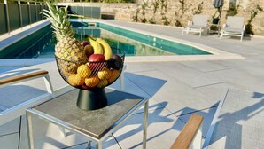 Un bol de fruita damunt una taula a prop d'una zona de piscina amb gandules i una paret de pedra al fons.