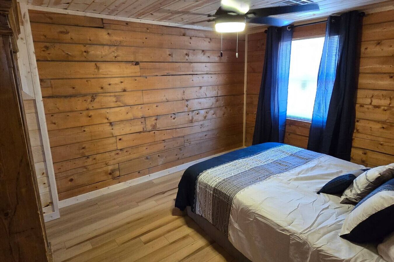 A wooden-paneled room with a bed, a ceiling fan, and a window with curtains.