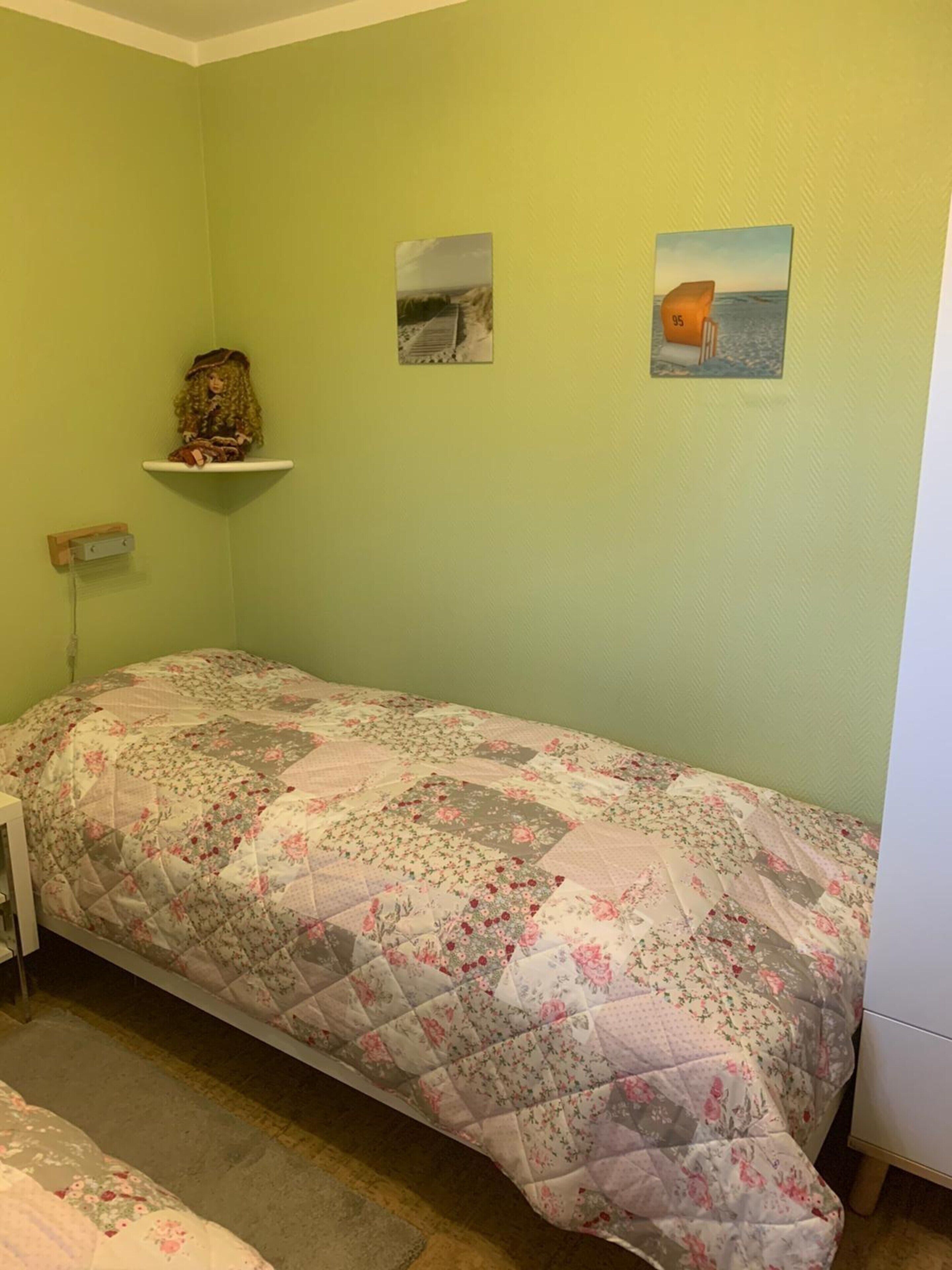 A room with a single bed, a small shelf with a doll, and two framed pictures on the wall.