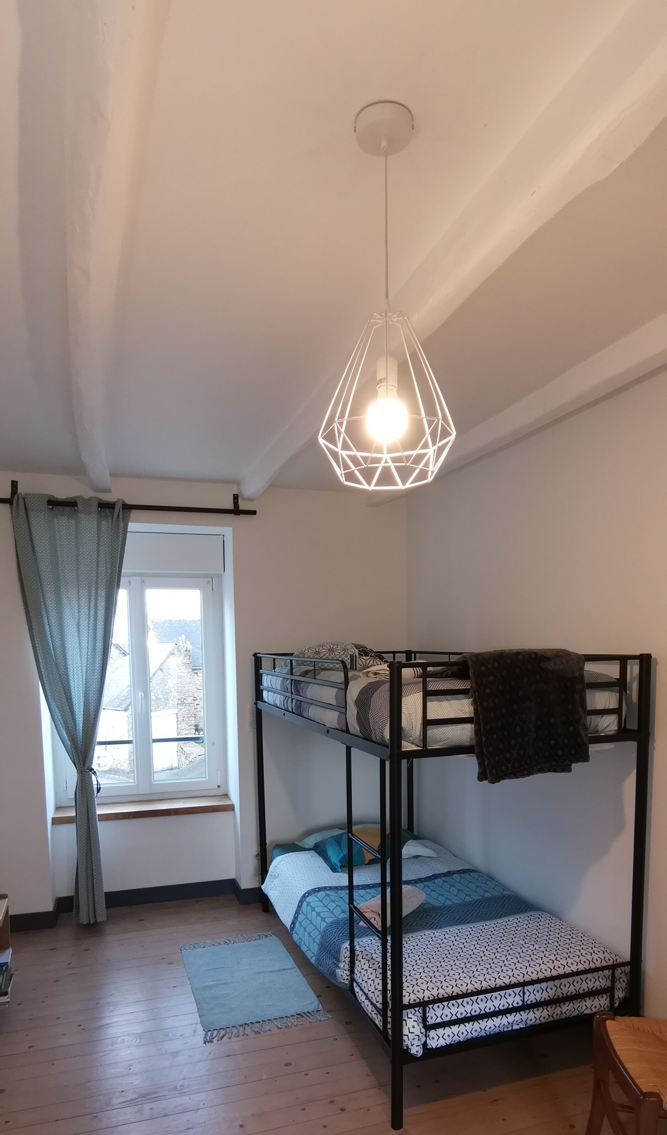 Een studentenkamer met een stapelbed, een hanglamp, een raam met gordijnen en uitzicht op buiten.