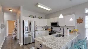 Una cocina moderna con armarios blancos, electrodomésticos de acero inoxidable, encimera de granito y taburetes de bar color turquesa.