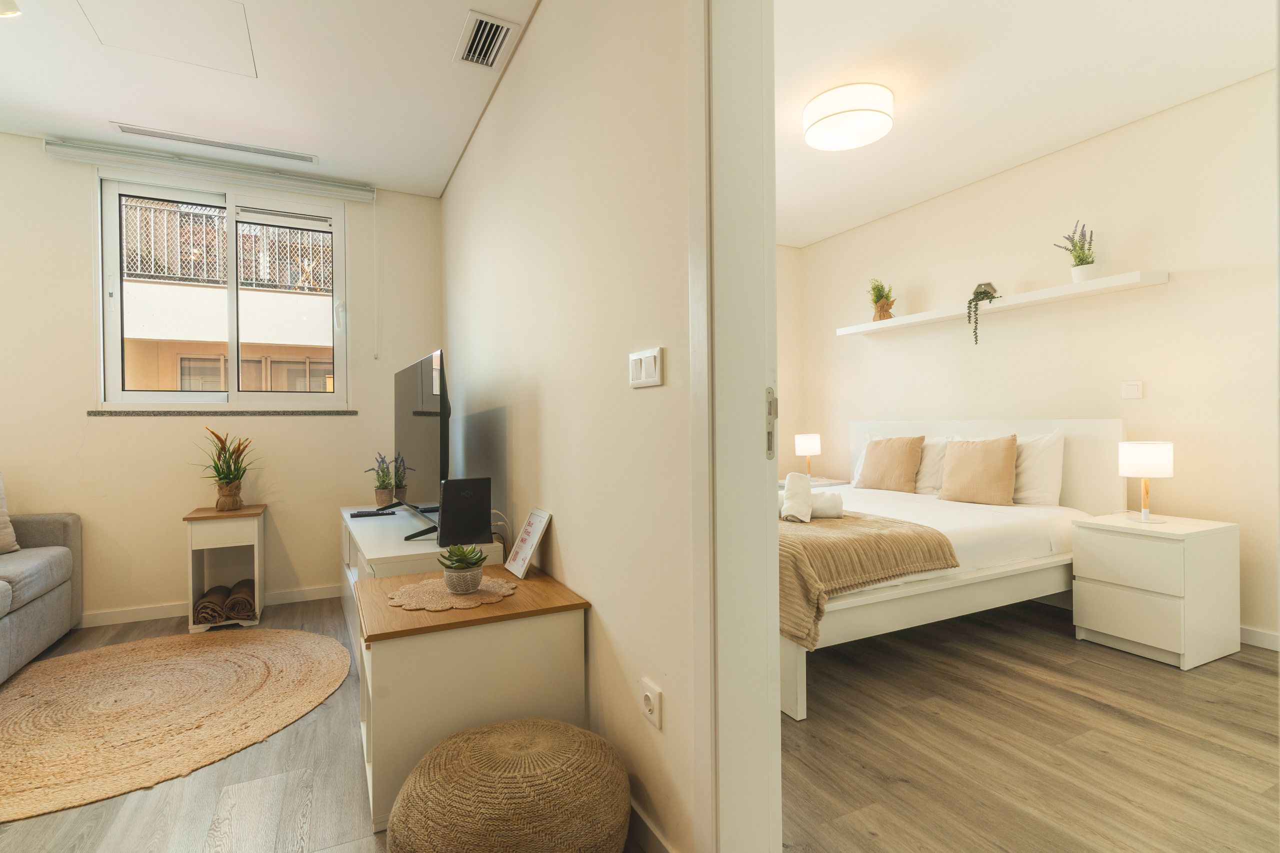 Un espace de vie compact comprenant un lit, des tables de chevet, un bureau avec un ordinateur, un petit coin salon et une fenêtre donnant sur un paysage agréable.