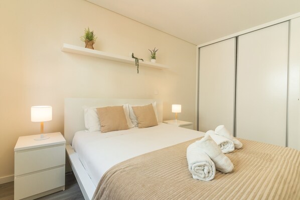 A hotel room with a bed, two bedside tables with lamps, a wardrobe, and a small shelf with plants.
