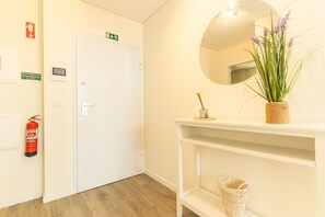 A hallway with a white door, emergency exit sign, fire extinguisher, and a mirror on a shelf with a plant. - Cassia Loft by Atlantic Holiday (Funchal)