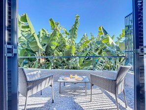 Un espacio exterior con asientos de mimbre y una mesa, rodeado de grandes plantas de plátano y un cielo azul despejado.