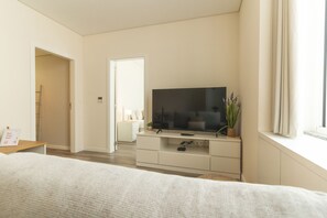 Habitación de hotel con cama, televisor sobre un mueble, mesita de noche, planta y ventana con cortinas.