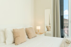Habitación de hotel con cama, almohadas, lámpara de noche, espejo y vistas al exterior.