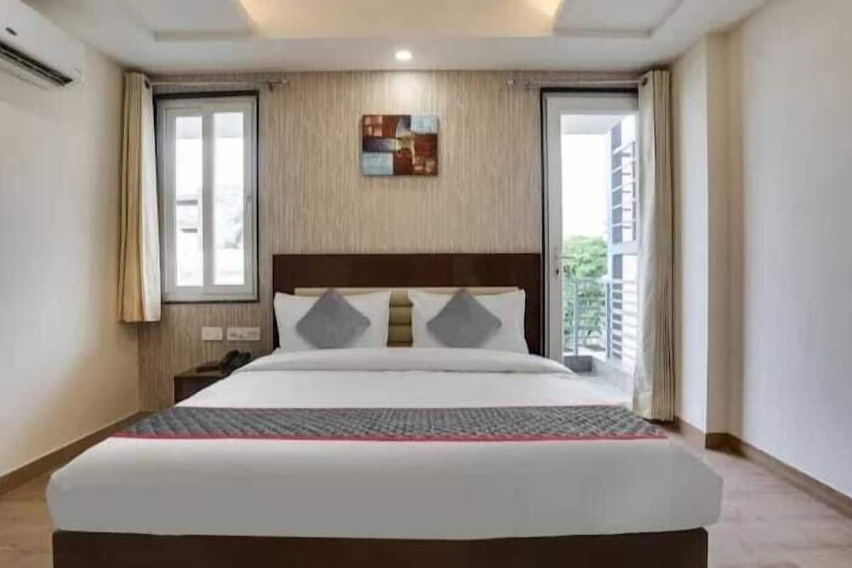 A hotel room with a large bed, a wooden headboard, two windows with curtains, a small bedside table, and a view of greenery outside.