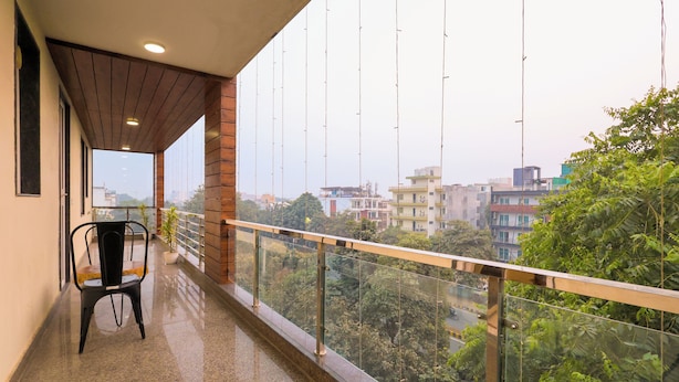A balcony with a glass railing, a chair, and a view of buildings and trees. - Nikko Palm Near Huda City Centre (Gurugram)