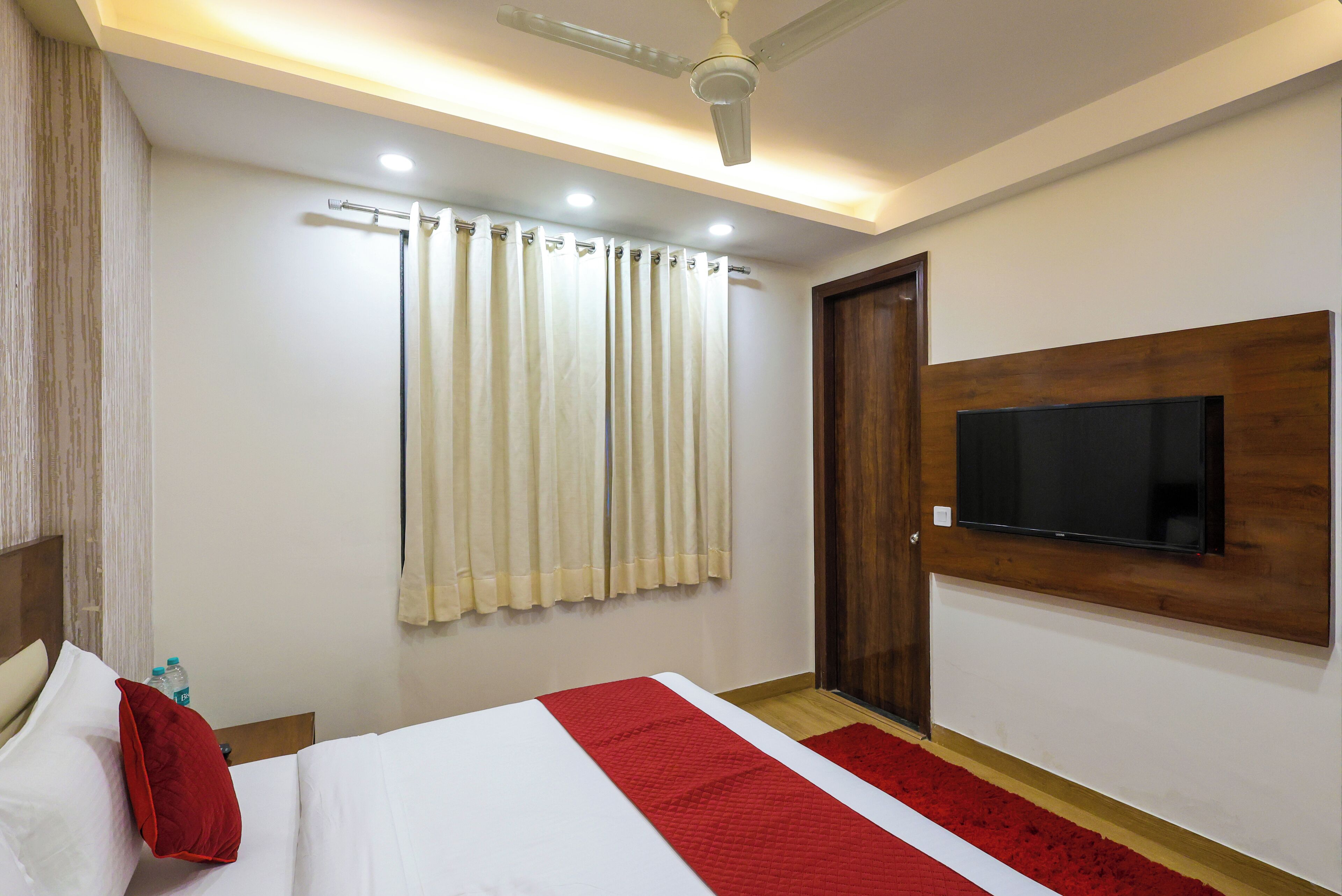 Une chambre d’hôtel avec un lit, un téléviseur fixé au mur, un ventilateur de plafond et une fenêtre avec des rideaux.