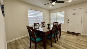 Un comedor con mesa y sillas de madera, un ventilador de techo y dos ventanas con persianas.