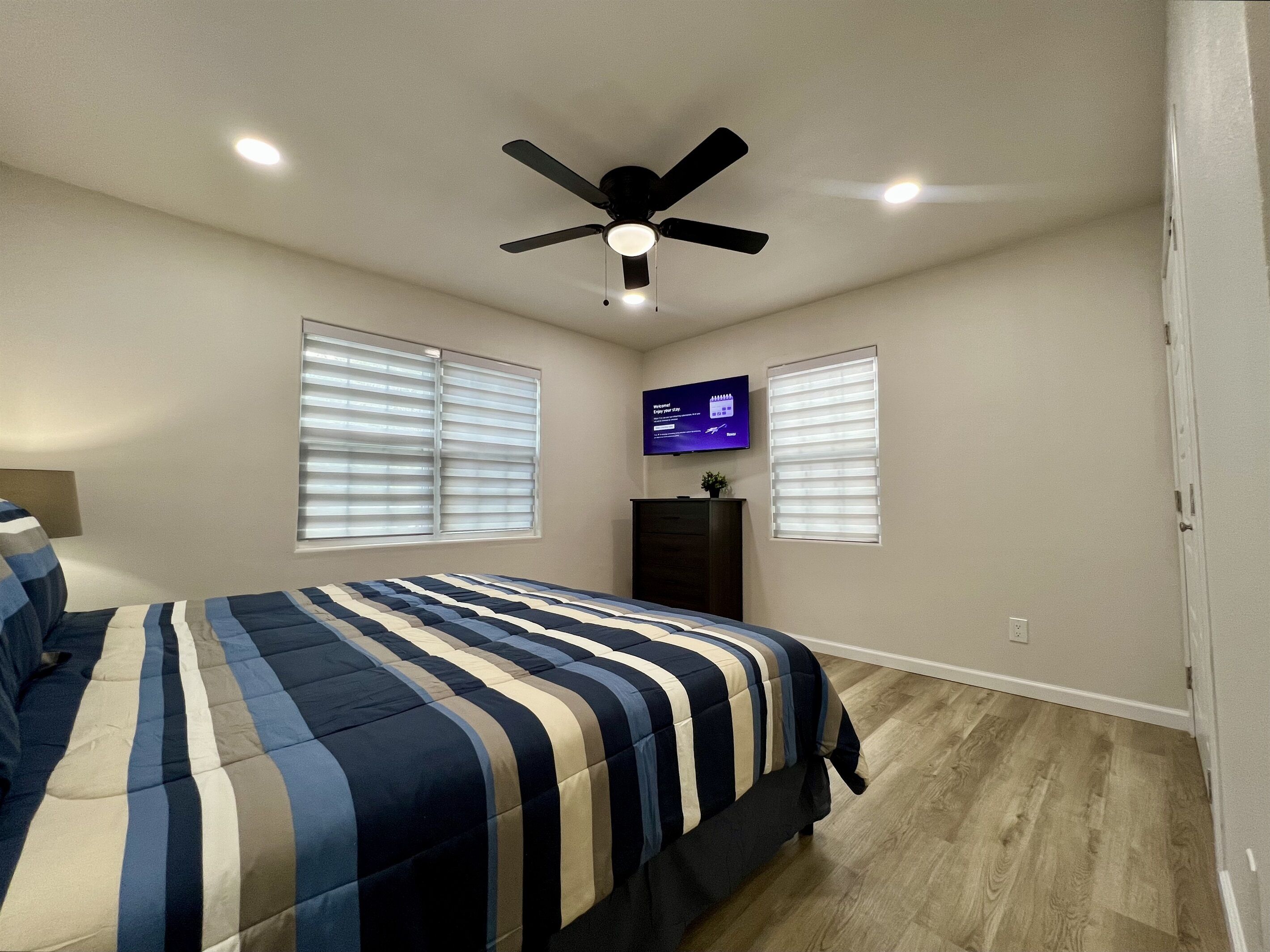Un dormitorio con cama, ventilador de techo, televisión y dos ventanas con persianas.