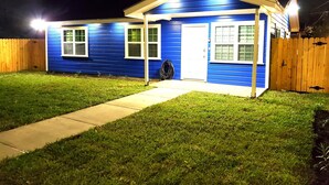 Una casa pequeña de un piso, con fachada azul y detalles blancos. Cuenta con un porche delantero con una puerta y dos ventanas. Una cerca de madera rodea la propiedad, y un camino de concreto lleva a la puerta principal.