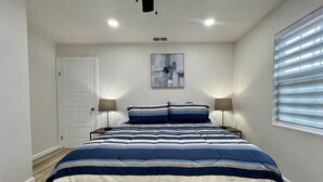 Un dormitorio con cama, dos mesitas de noche, una puerta, una ventana con persianas y una lámpara en el techo.