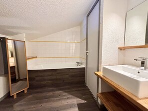 A modern bathroom with a wooden floor, white tiles, a bathtub, and a large mirror.