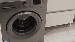 A compact, gray front-loading washing machine in a bathroom with white marble tiles and a white sink. - 2 rooms 37M2 4p.tout neuf.limite Paris12e.30mn DISNEY, 9mn CHAMPS ELYSEE+LOUVRE  (Vincennes)
