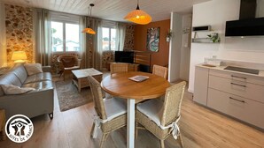 A modern living room with a sofa, a dining table with chairs, a TV, and a kitchen area with cabinets and a microwave. - Modern house with AC, garden and parking, near Noirmoutier beach (Barbâtre)