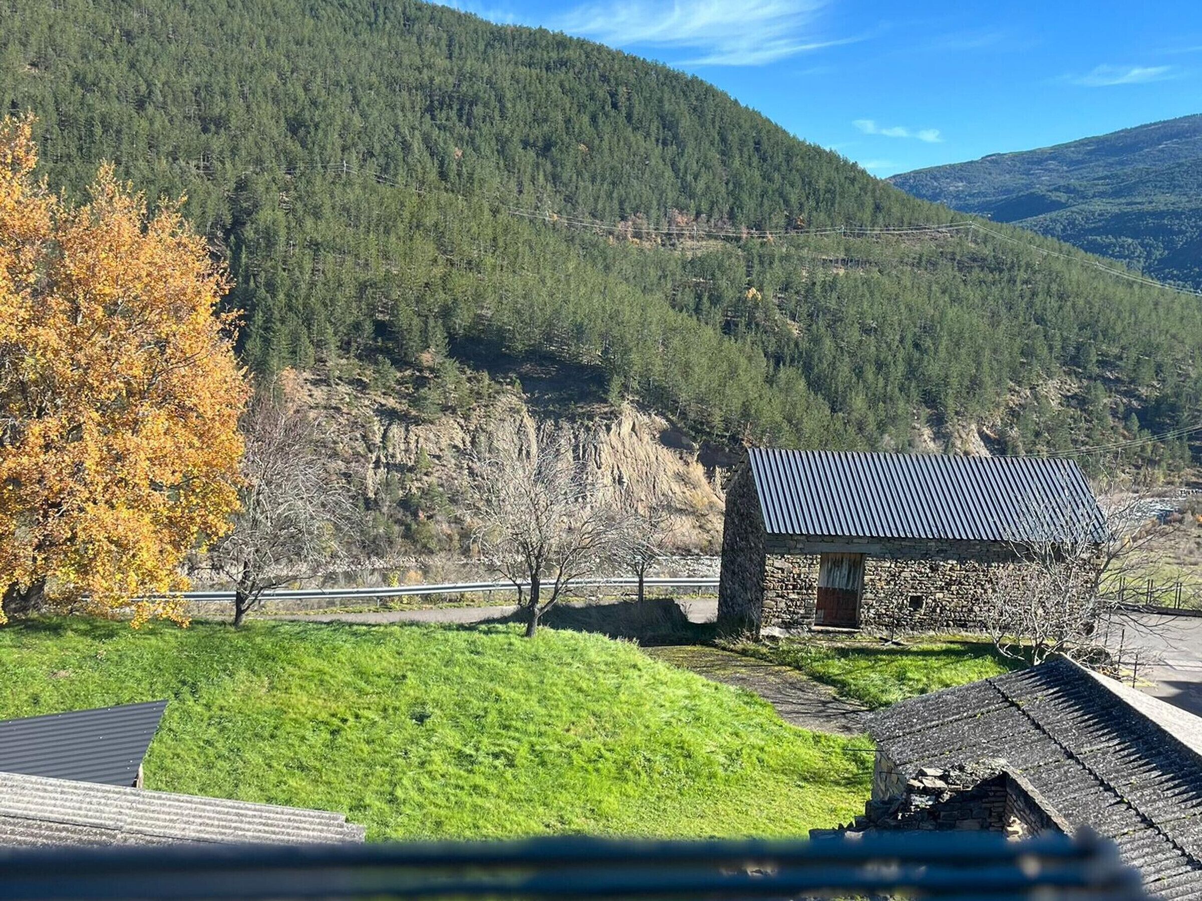 A stone building with a metal roof, surrounded by a grassy area and a forested mountain backdrop.