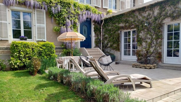 Un patio con mobiliario exterior, una puerta azul y un jardín con diversas plantas.
