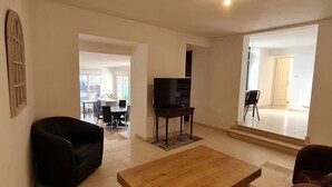 Un salón espacioso con una mesa de centro de madera, sillones negros y un televisor sobre un mueble bajo. Grandes ventanales ofrecen luz natural y vistas al exterior.