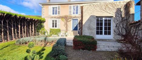 Une maison à deux étages avec une façade en pierre, un petit jardin agrémenté d’un chemin de gravier et une porte bleue.