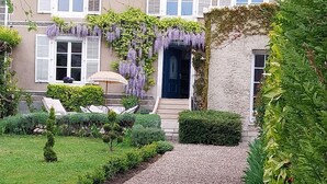 Un sendero en un jardín que conduce a un edificio con puerta azul, rodeado de follaje y una pequeña zona de estar con una mesa y sillas.