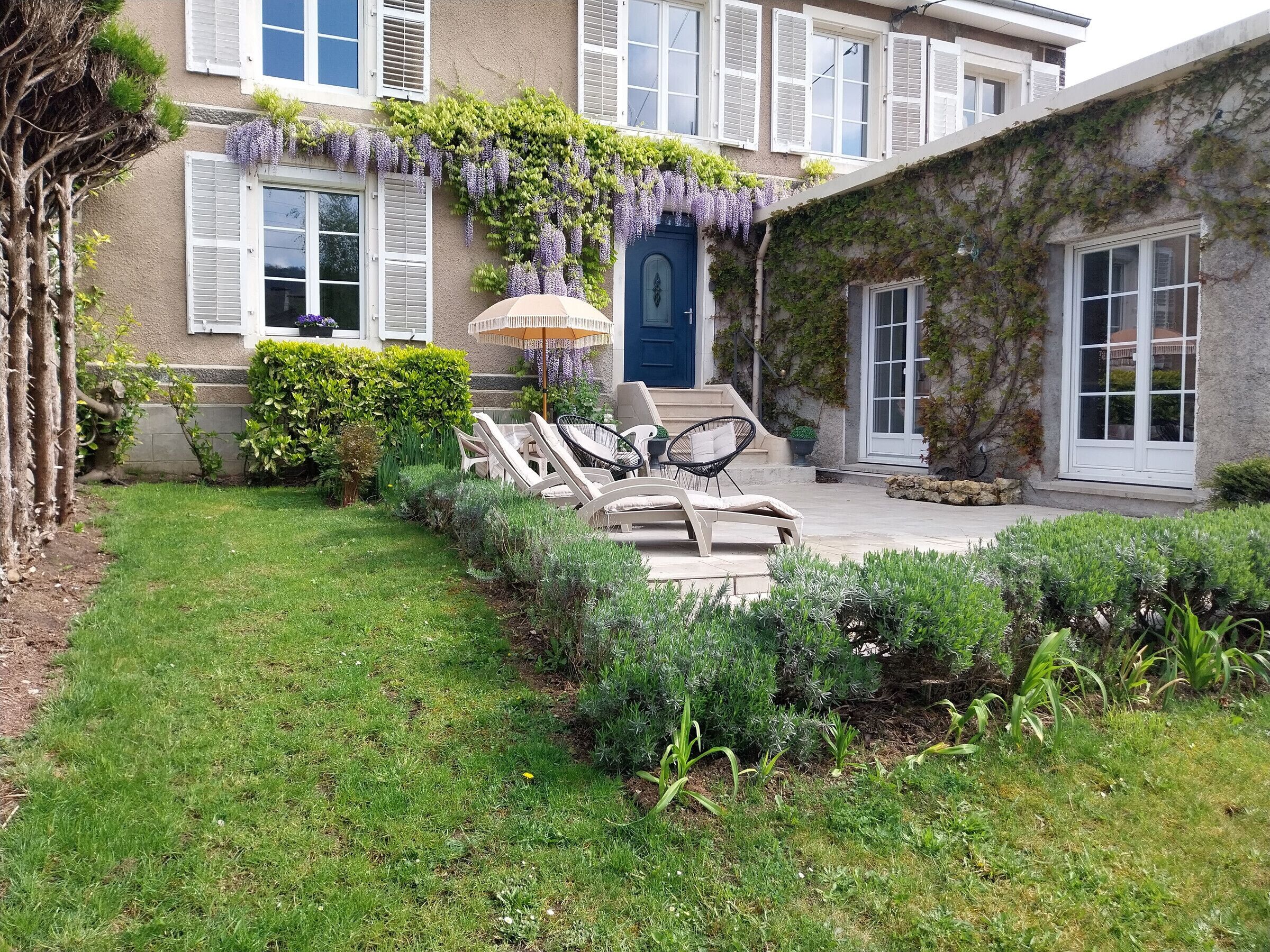 A courtyard with a blue door, white shutters, and a pergola with purple flowers. There are lounge chairs and a table under the pergola.