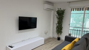 Una sala de estar moderna con un televisor de pantalla plana montado en la pared, un mueble de entretenimiento blanco, un sofá gris con un cojín amarillo, una planta verde alta y un ventanal con vista a edificios.