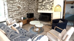 A living room with a stone wall, a fireplace, a television, a sofa, a chair, and a small round table. - Spacious house with garden in the heart of Lomener (Groix)