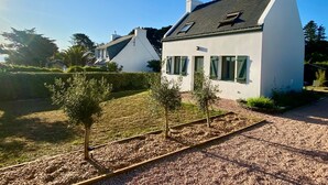A modern house with a sloped roof, white exterior walls, and a small garden area with a gravel path and young trees. - Charming house near beach in Groix, 2 bedrooms (Groix)