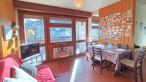 Quarto acolhedor com um sofá vermelho, paredes de madeira, uma mesa de jantar posta para quatro pessoas e uma parede decorada com fotografias.