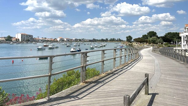 A waterfront promenade with a wooden walkway, boats in the water, and buildings in the background.