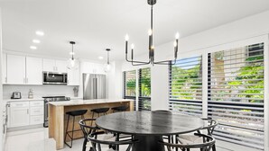 Una cocina moderna con armarios blancos, una isla central con taburetes, una mesa redonda para comer y un ventanal con persianas.