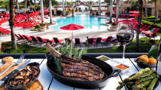 Una mesa de comedor con un filete a la parrilla, espárragos y una copa de vino tinto, con vistas a una zona de piscina con sombrillas y sillones de descanso rojos.