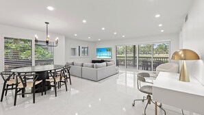 Una sala de estar moderna con comedor, un conjunto de sofás, un televisor y un amplio ventanal con persianas.