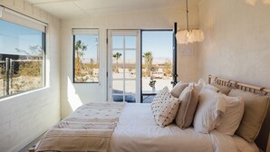 Un dormitorio con cama, almohadas, una mesita de noche y vista a un paisaje desértico a través de una puerta corrediza.