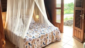 Ein Zimmer mit einem Bett unter einem Baldachin, einer zur Außenwelt führenden Tür und Blick ins Grüne.